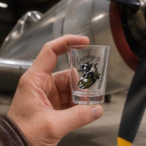 May include: A clear glass shot glass featuring a black and white cartoon cat wearing goggles and a scarf, riding an olive green bomb with a shark mouth graphic. The text Lucky 13 is printed in black script above the cat illustration.
