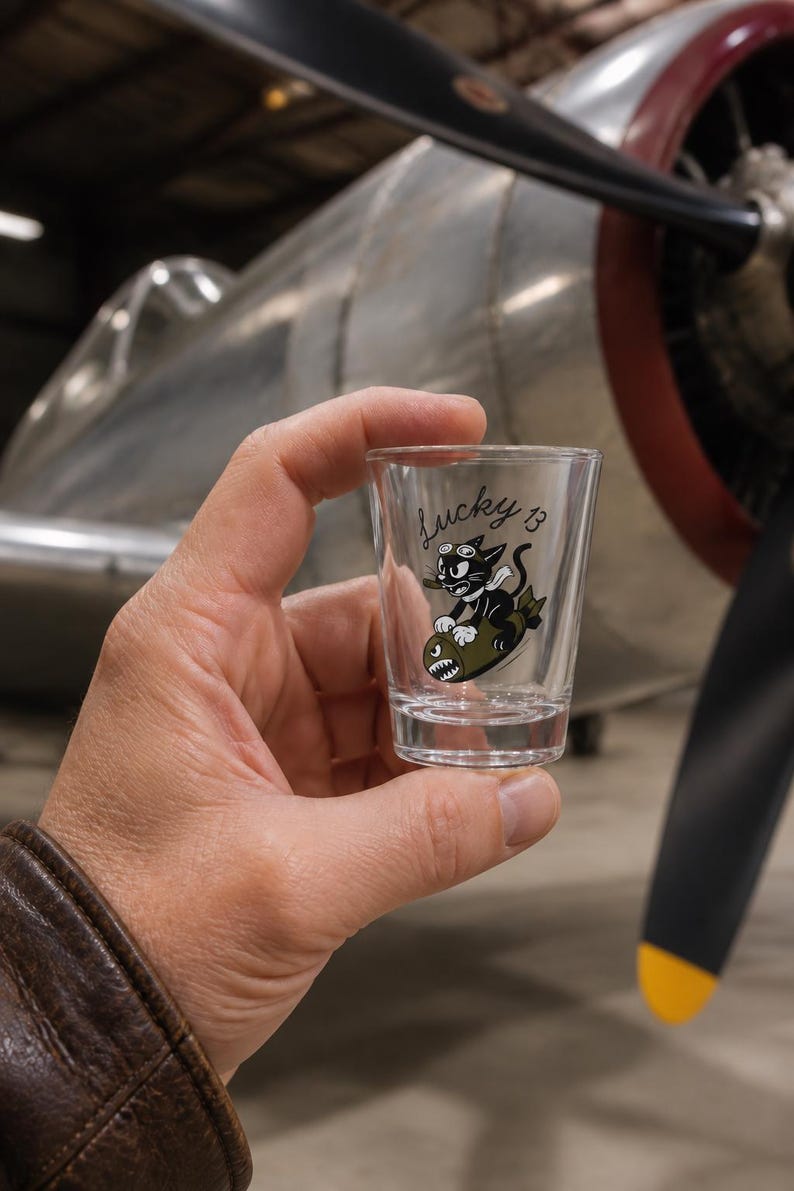 May include: A clear glass shot glass featuring a black and white cartoon cat wearing goggles and a scarf, riding an olive green bomb with a shark mouth graphic. The text Lucky 13 is printed in black script above the cat illustration.