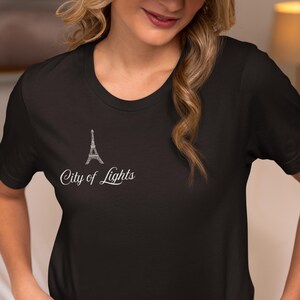 May include: Black t-shirt featuring a white Eiffel Tower design and the text "City of Lights." The shirt has a classic crew neck and short sleeves. The design is located on the upper left chest area.