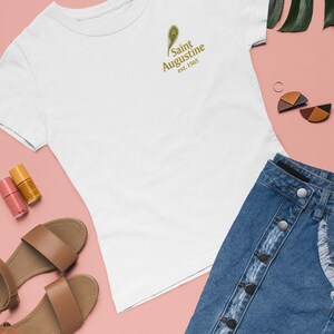 May include: White t-shirt with a gold peacock feather and text that reads "Saint Augustine est. 1565". The shirt is paired with a denim skirt, brown sandals, and nail polish on a pink background.