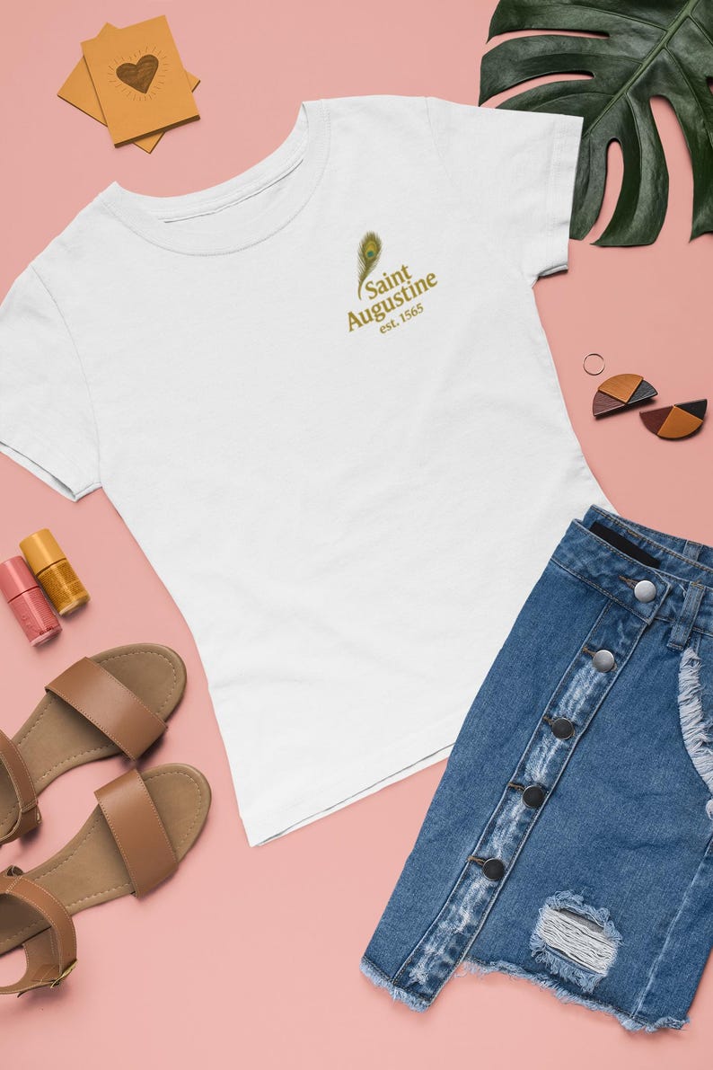 May include: White t-shirt with a gold peacock feather and text that reads "Saint Augustine est. 1565". The shirt is paired with a denim skirt, brown sandals, and nail polish on a pink background.