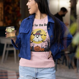 May include: A light pink t-shirt with a Halloween-themed graphic. The design features a ghost wearing a witch hat in front of a yellow moon, with bats, pumpkins, and trees. The shirt is worn with a blue corduroy jacket and denim shorts.