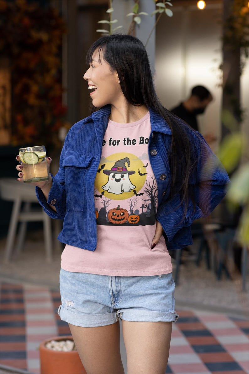 May include: A light pink t-shirt with a Halloween-themed graphic. The design features a ghost wearing a witch hat in front of a yellow moon, with bats, pumpkins, and trees. The shirt is worn with a blue corduroy jacket and denim shorts.