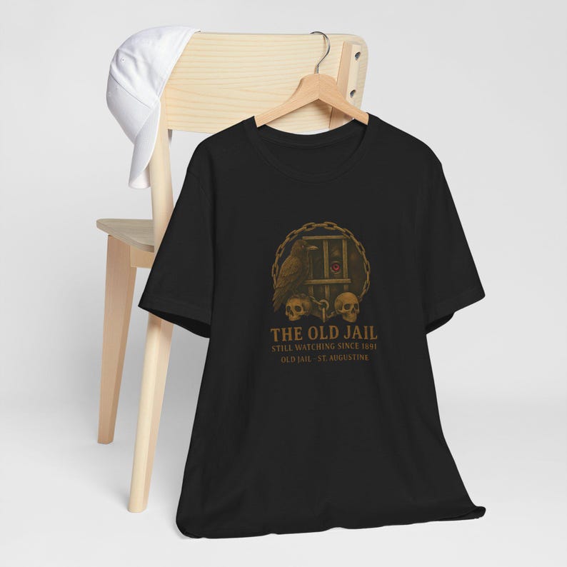 May include: Black t-shirt with a graphic design featuring a raven, skulls, and a jail cell. The design is in shades of brown and gold, with the words 'THE OLD JAIL' and 'STILL WATCHING SINCE 1891 OLD JAIL - ST. AUGUSTINE' printed below. The t-shirt is displayed on a hanger against a light background.