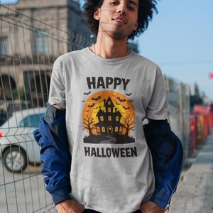 May include: A light gray t-shirt with a Halloween graphic. The graphic features a haunted house silhouette in black against an orange moon, with the words "HAPPY HALLOWEEN" in black above and below the image.
