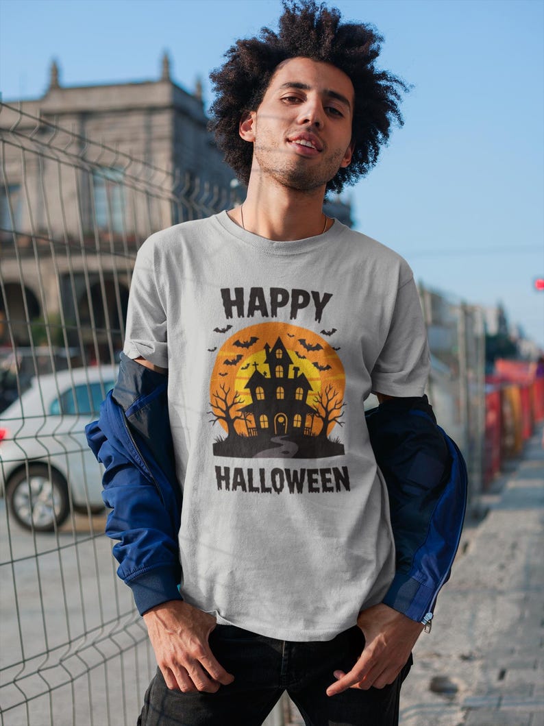 May include: A light gray t-shirt with a Halloween graphic. The graphic features a haunted house silhouette in black against an orange moon, with the words "HAPPY HALLOWEEN" in black above and below the image.