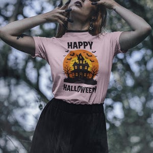 May include: A light pink t-shirt with a Halloween graphic. The graphic features a haunted house, bats, and the words "HAPPY HALLOWEEN". The person is wearing a black skirt.
