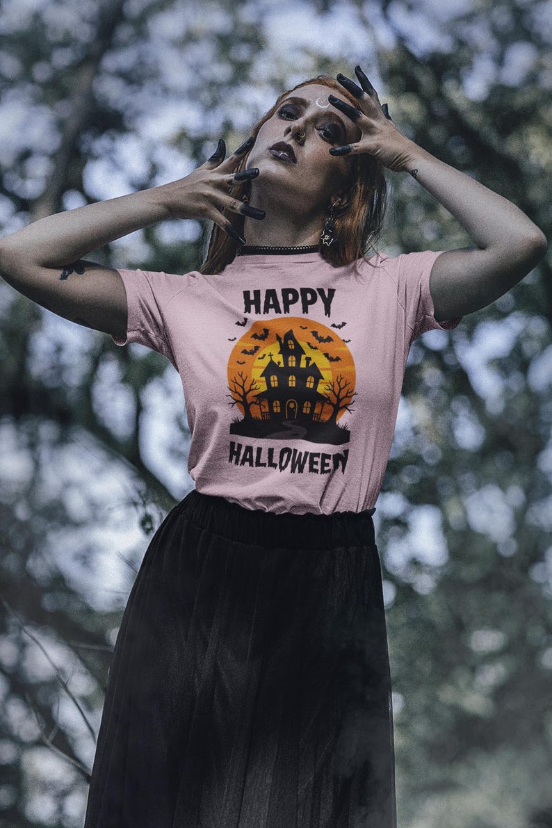 May include: A light pink t-shirt with a Halloween graphic. The graphic features a haunted house, bats, and the words "HAPPY HALLOWEEN". The person is wearing a black skirt.