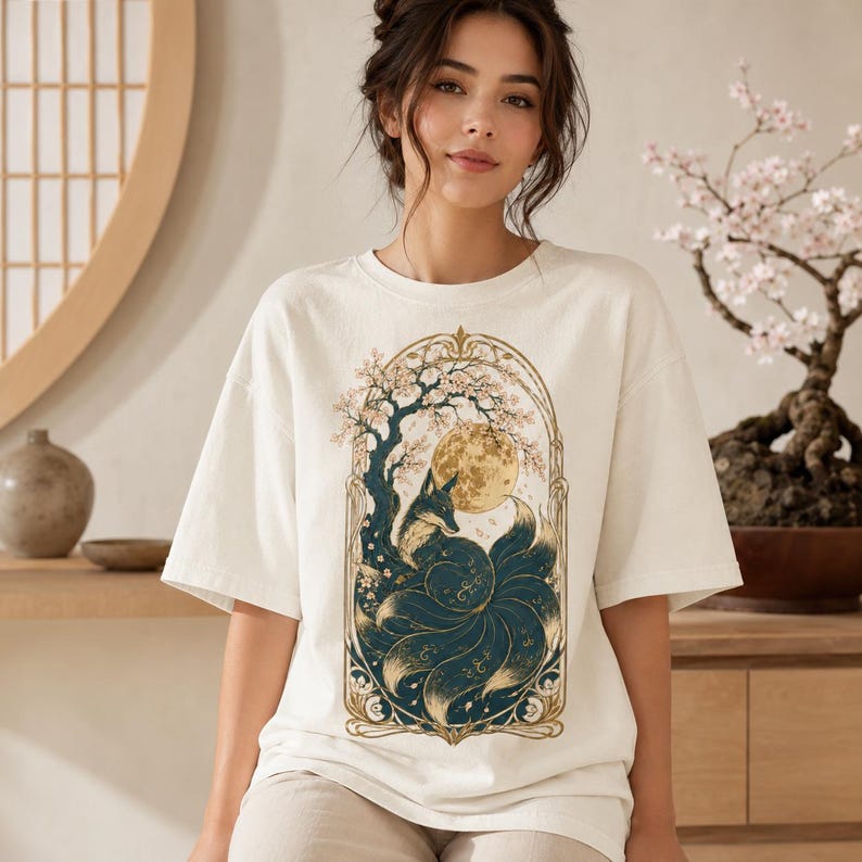 May include: An off-white short-sleeve t-shirt featuring a central graphic print of a multi-tailed fox sitting under a large moon and a flowering tree, all enclosed within an ornate gold-coloured archway frame with intricate decorative details.