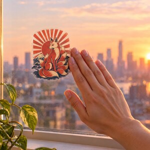 May include: A clear adhesive sticker featuring a multi-tailed fox sitting in front of a red sunburst pattern, a mountain silhouette, and small pine trees. The sticker is applied to a glass window pane with a city skyline visible in the background.