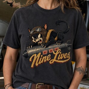 以下が含まれることがあります： チャコールグレーのTシャツ。茶色の飛行帽とゴーグルを身につけ、灰色の爆弾にまたがった黒猫のグラフィックが描かれています。爆弾の下には黄色の筆記体で「Nine Lives」という文字と、3つの小さな赤い太陽のアイコンがプリントされています。