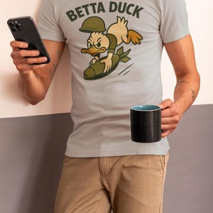May include: Pale grey t-shirt featuring the text "Betta Duck" and a cartoon duck riding a bomb. The duck wears a helmet and olive green clothing. The person holds a black mug and a smartphone.