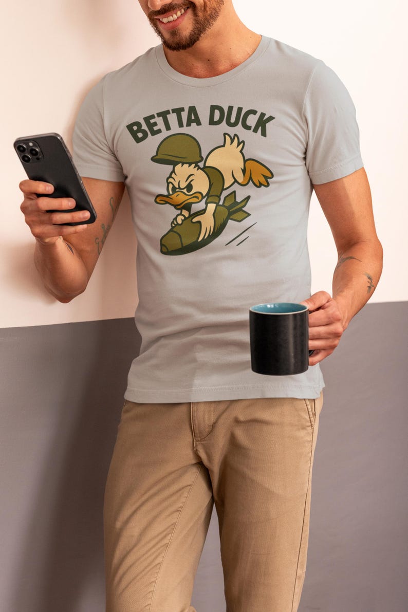 May include: Pale grey t-shirt featuring the text "Betta Duck" and a cartoon duck riding a bomb. The duck wears a helmet and olive green clothing. The person holds a black mug and a smartphone.