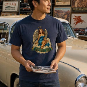 May include: A navy blue short-sleeve t-shirt featuring a graphic print of a winged figure with auburn hair wearing a blue dress, seated on clouds with a halo above, and a banner below with the text HEAVENLY BODY in gold capital letters.