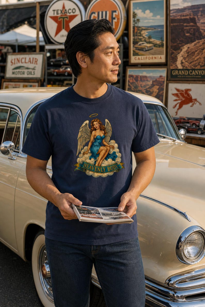 May include: A navy blue short-sleeve t-shirt featuring a graphic print of a winged figure with auburn hair wearing a blue dress, seated on clouds with a halo above, and a banner below with the text HEAVENLY BODY in gold capital letters.