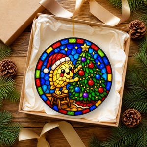 Puede incluir: Un adorno circular estilo vidriera que muestra un personaje de masa amarilla con un gorro de Pap&aacute; Noel rojo, de pie sobre un peque&ntilde;o taburete de madera decorando un &aacute;rbol de Navidad verde con adornos rojos y azules sobre un fondo azul.