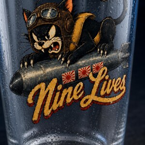 Op de afbeelding: Een helder glazen beker met een afbeelding van een zwarte kat met een bruine vliegeniershelm en bril, rijdend op een grijze bom. De bom toont drie rood-witte zonne-emblemen. De tekst Nine Lives is in het geel en rood op het glas gedrukt.
