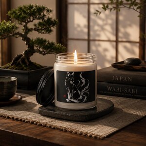 May include: A clear glass jar candle with a lit wick and a black lid resting beside it. The label features a white ink illustration of a multi-tailed fox on a black background. The candle sits on a dark stone coaster on a woven fabric mat.