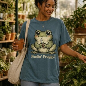 Może przedstawiać: Niebieski t-shirt z kr&oacute;tkim rękawem z grafiką zielonej żaby trzymającej mały n&oacute;ż oraz napisem "Feelin' Froggy!" pod ilustracją. Koszulka ma okrągły dekolt, luźny kr&oacute;j i jest zestawiona z beżową torbą pł&oacute;cienną typu tote.