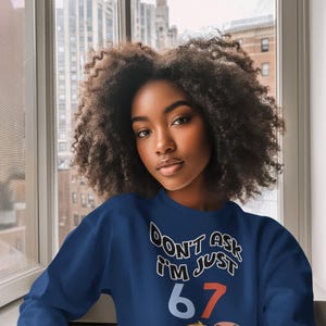 May include: A navy blue sweatshirt with the text "DON'T ASK I'M JUST 67" in white and red. A cartoon chick with the word "meh" is below the text. The sweatshirt is worn by a person with dark curly hair.