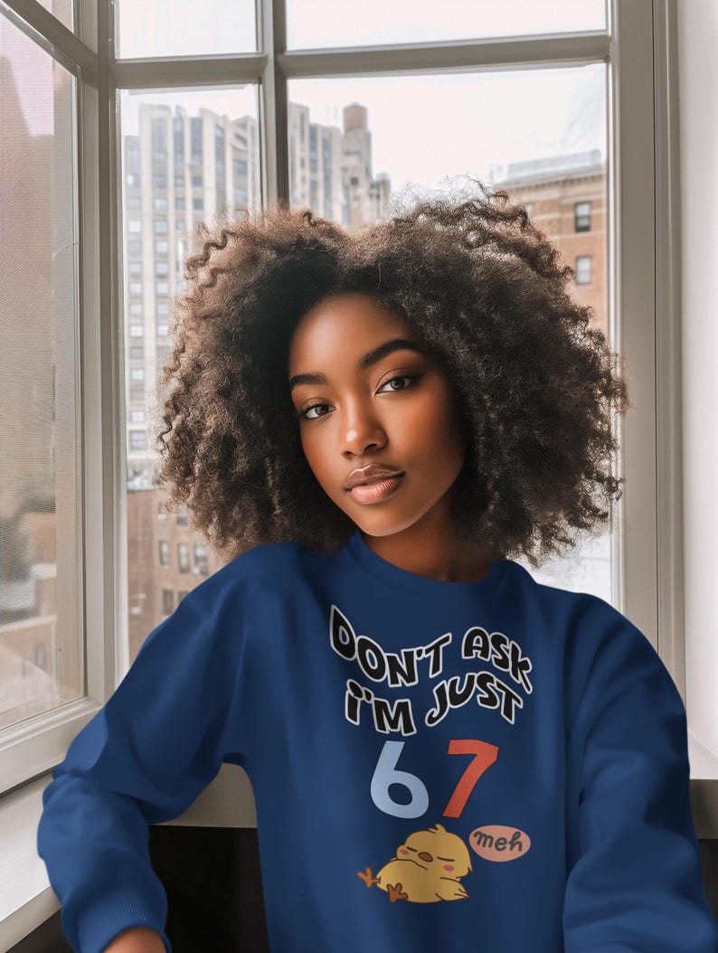 May include: A navy blue sweatshirt with the text "DON'T ASK I'M JUST 67" in white and red. A cartoon chick with the word "meh" is below the text. The sweatshirt is worn by a person with dark curly hair.