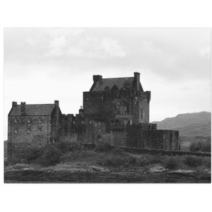 May include: Black and white photograph of a medieval castle with multiple towers and a stone wall. The castle is situated on a grassy area with a mountain range in the background under a cloudy sky.