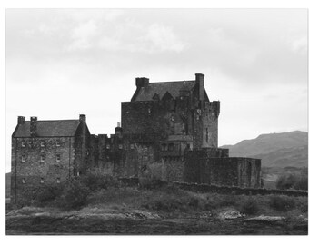 May include: Monochrome image of a historic castle with several turrets and a stone wall. The castle is set on a grassy bank, with a mountain range in the distance, under an overcast sky.