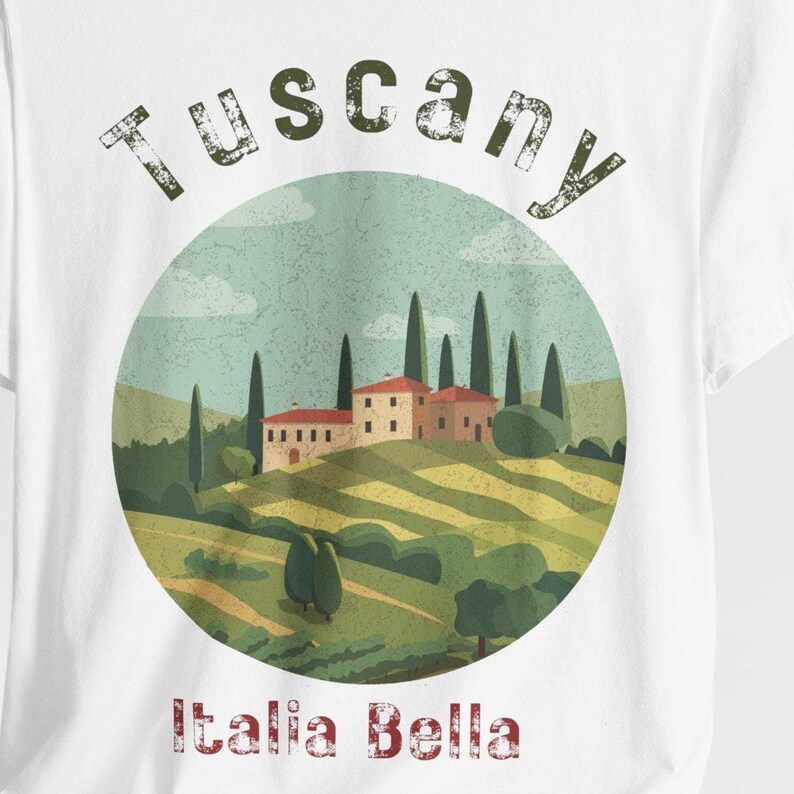 May include: White t-shirt featuring a circular graphic of a Tuscan landscape with rolling hills, cypress trees, and buildings. The word "Tuscany" arches across the top, and "Italia Bella" is printed at the bottom.