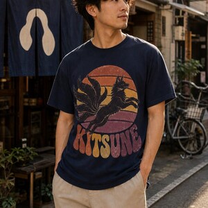 May include: A navy blue short-sleeve t-shirt featuring a graphic print of a black nine-tailed fox leaping in front of a sunset circle. The word KITSUNE is printed in a bold, retro-style font below the fox, with a distressed texture throughout.
