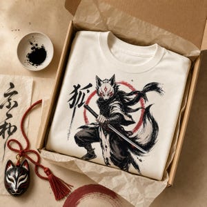 May include: A cream-colored t-shirt featuring a graphic of a fox-masked warrior holding a katana, with a red circle and the Japanese kanji for fox. The shirt is inside a cardboard box with a small black and white fox mask pendant nearby.