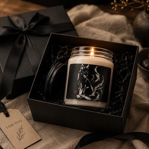 May include: A scented candle in a clear glass jar with a black label featuring a white fox illustration, placed inside a black gift box with a black ribbon. A brown paper gift tag with the text FOR YOU and a leaf graphic is attached to the ribbon.