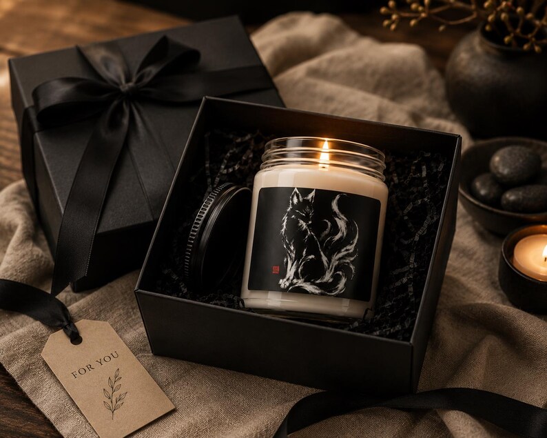 May include: A scented candle in a clear glass jar with a black label featuring a white fox illustration, placed inside a black gift box with a black ribbon. A brown paper gift tag with the text FOR YOU and a leaf graphic is attached to the ribbon.