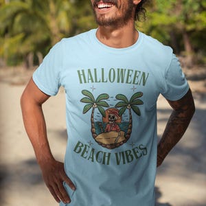May include: Pale blue t-shirt with the text "HALLOWEEN BEACH VIBES" in green. The design includes a cartoon skeleton on a beach with palm trees. The person in the image is wearing sunglasses.