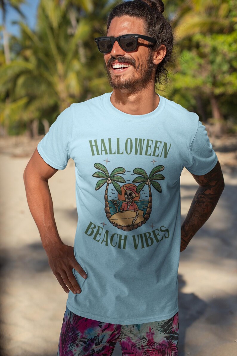 May include: Pale blue t-shirt with the text "HALLOWEEN BEACH VIBES" in green. The design includes a cartoon skeleton on a beach with palm trees. The person in the image is wearing sunglasses.