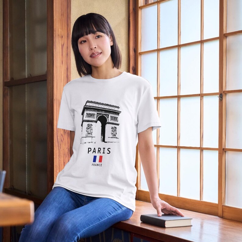 May include: White t-shirt featuring a black graphic of the Arc de Triomphe in Paris. Below the graphic, the word 'PARIS' is printed, with the French flag underneath. The shirt is worn by a person sitting near a window.