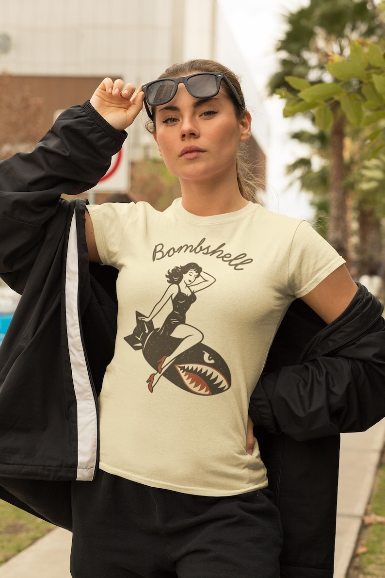 May include: Cream-colored t-shirt featuring a vintage-style graphic of a woman riding a bomb, with the word "Bombshell" above. The design includes a woman in a black dress and red heels. The shirt is short-sleeved.