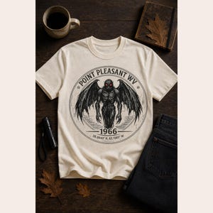 May include: A cream-colored short-sleeve t-shirt featuring a circular black graphic of a winged creature with glowing red eyes. The text reads Point Pleasant WV, 1966, and the coordinates 38.8443 N, 82.1385 W within the circular design.