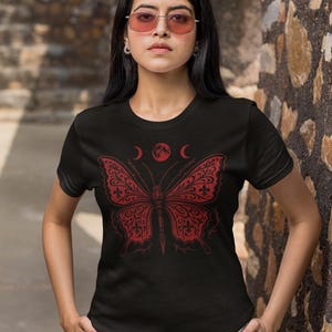 May include: Black t-shirt featuring a red butterfly design with crescent moons and a full moon above it. The butterfly has intricate patterns. The shirt is worn by a person with pink-tinted sunglasses.