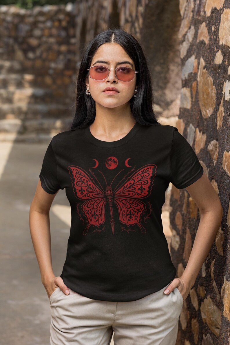 May include: Black t-shirt featuring a red butterfly design with crescent moons and a full moon above it. The butterfly has intricate patterns. The shirt is worn by a person with pink-tinted sunglasses.