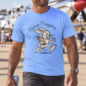 Puede incluir: Una camiseta de manga corta de color azul claro con un gr&aacute;fico de un conejo de dibujos animados corriendo mientras sostiene una bomba. Sobre el conejo, el texto "Hare Trigger" aparece en una fuente oscura y curva en el pecho.
