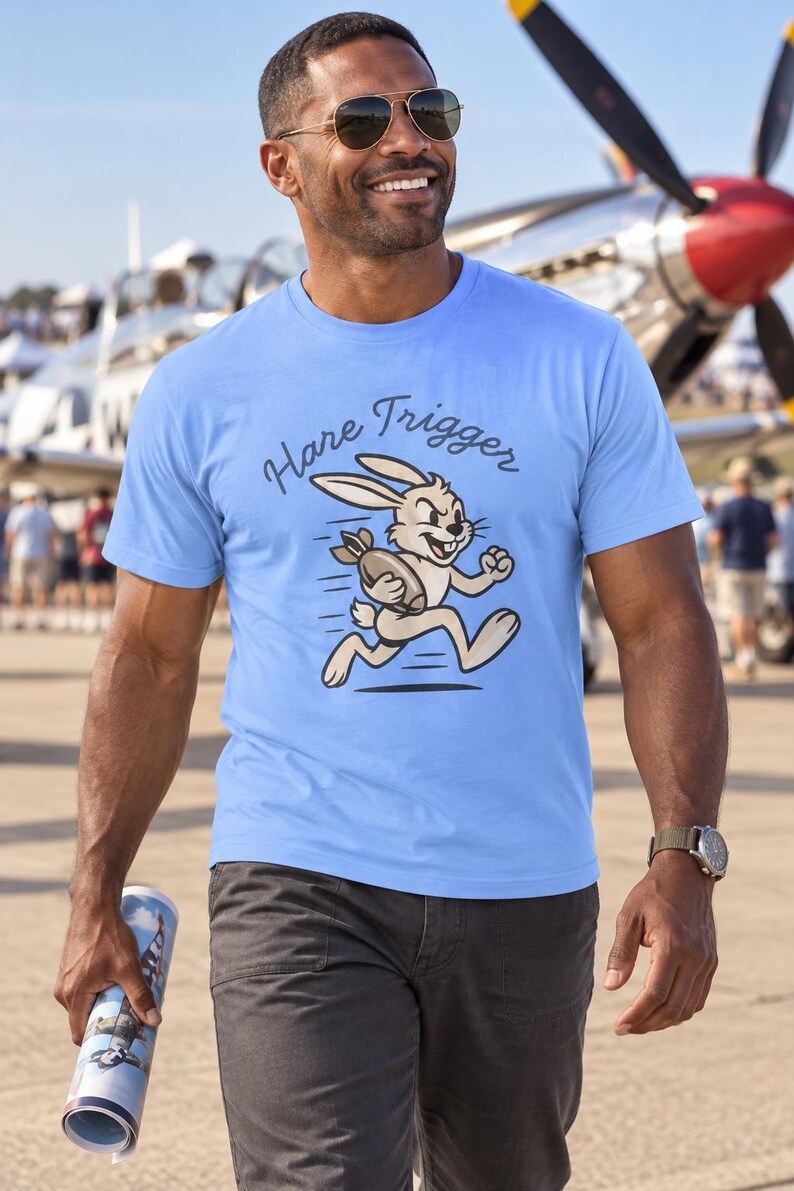 Puede incluir: Una camiseta de manga corta de color azul claro con un gr&aacute;fico de un conejo de dibujos animados corriendo mientras sostiene una bomba. Sobre el conejo, el texto "Hare Trigger" aparece en una fuente oscura y curva en el pecho.