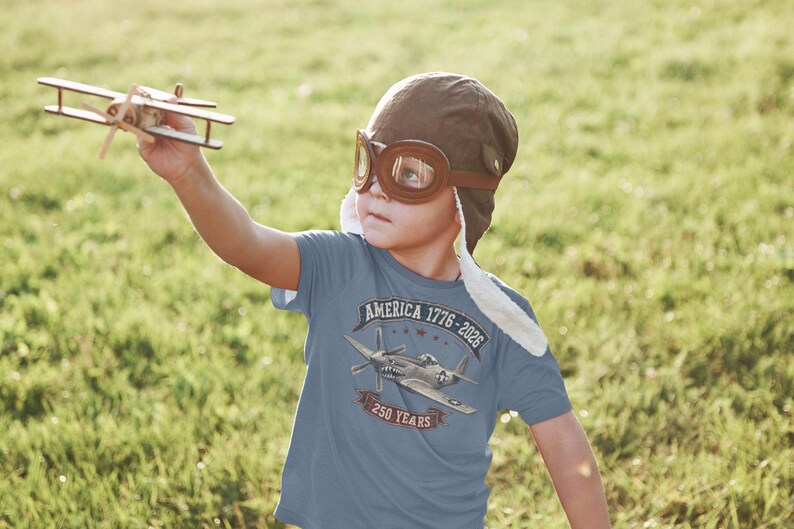 May include: A blue t-shirt featuring a graphic of a vintage fighter plane with the text AMERICA 1776-2026 and 250 YEARS. The shirt is worn with a brown aviator cap with goggles, and a small wooden toy airplane is held in the air.