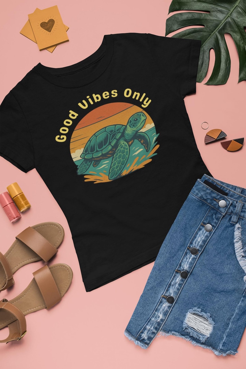 May include: Black t-shirt featuring a sea turtle graphic against a sunset backdrop, with the text "Good Vibes Only" in yellow. The image includes a denim skirt and sandals, set against a pink background.