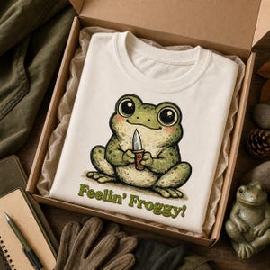 Może przedstawiać: Kremowy t-shirt z grafiką przedstawiającą zieloną żabę z dużymi oczami trzymającą mały n&oacute;ż. Pod żabą znajduje się zielony napis "Feelin' Froggy!". Koszulka jest złożona w kartonowym pudełku wyłożonym pogniecionym papierem pakowym.