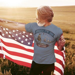 May include: A slate blue short-sleeved t-shirt featuring a graphic print with the text AMERICA 1776-2026 and 250 YEARS. The design includes a historical figure standing on a shore with ships in the distance, all rendered in muted earth tones.