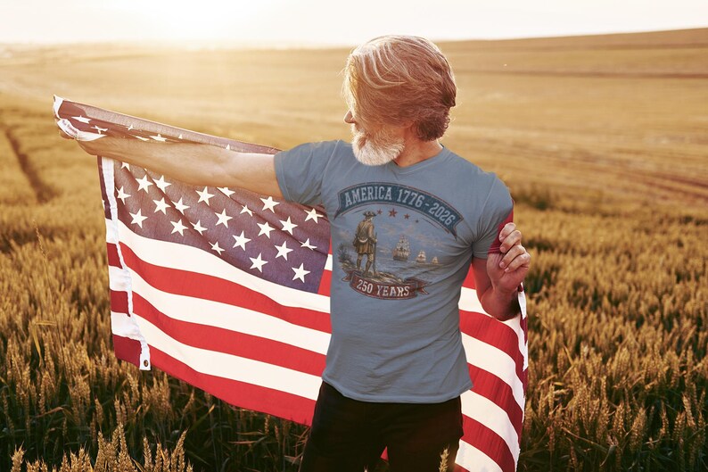 May include: A slate blue short-sleeved t-shirt featuring a graphic print with the text AMERICA 1776-2026 and 250 YEARS. The design includes a historical figure standing on a shore with ships in the distance, all rendered in muted earth tones.