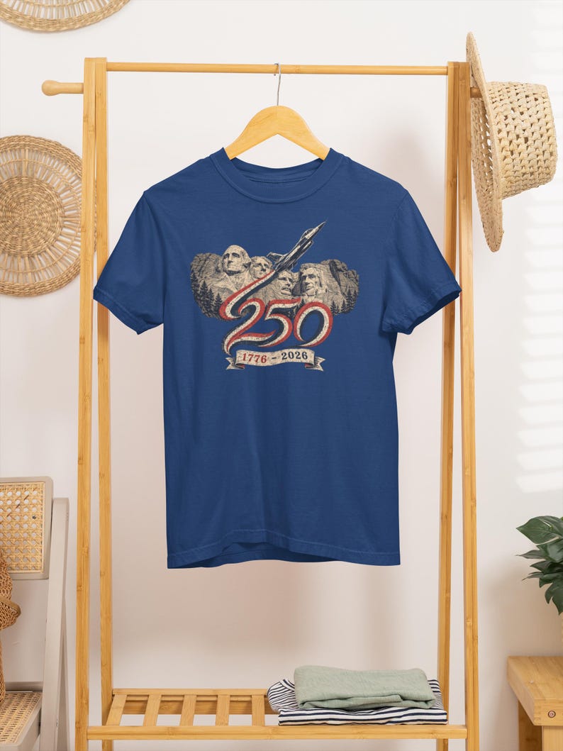 May include: A navy blue t-shirt featuring a graphic of Mount Rushmore with a jet flying above. The design includes the large number 250 in red and white, with a banner below displaying the text 1776 - 2026 in a vintage, distressed style.