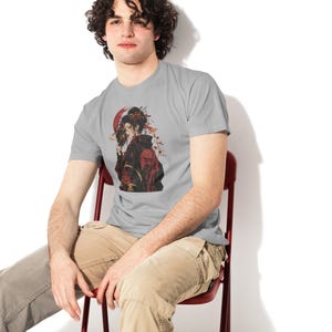 May include: A light gray short-sleeved t-shirt featuring a graphic illustration of a person in a black and red kimono holding a mask. The design includes a large red circle in the background, delicate floral accents, and traditional architectural motifs.