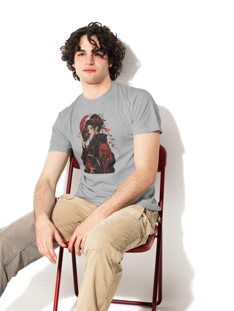 May include: A light gray short-sleeved t-shirt featuring a graphic illustration of a person in a black and red kimono holding a mask. The design includes a large red circle in the background, delicate floral accents, and traditional architectural motifs.