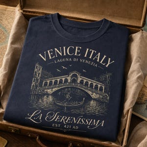 May include: A navy blue t-shirt featuring a cream-colored graphic of the Rialto Bridge in Venice, Italy. The text reads VENICE ITALY, LAGUNA DI VENEZIA, LA SERENISSIMA, and EST. 421 AD. The design includes a gondola, birds, and architectural details.
